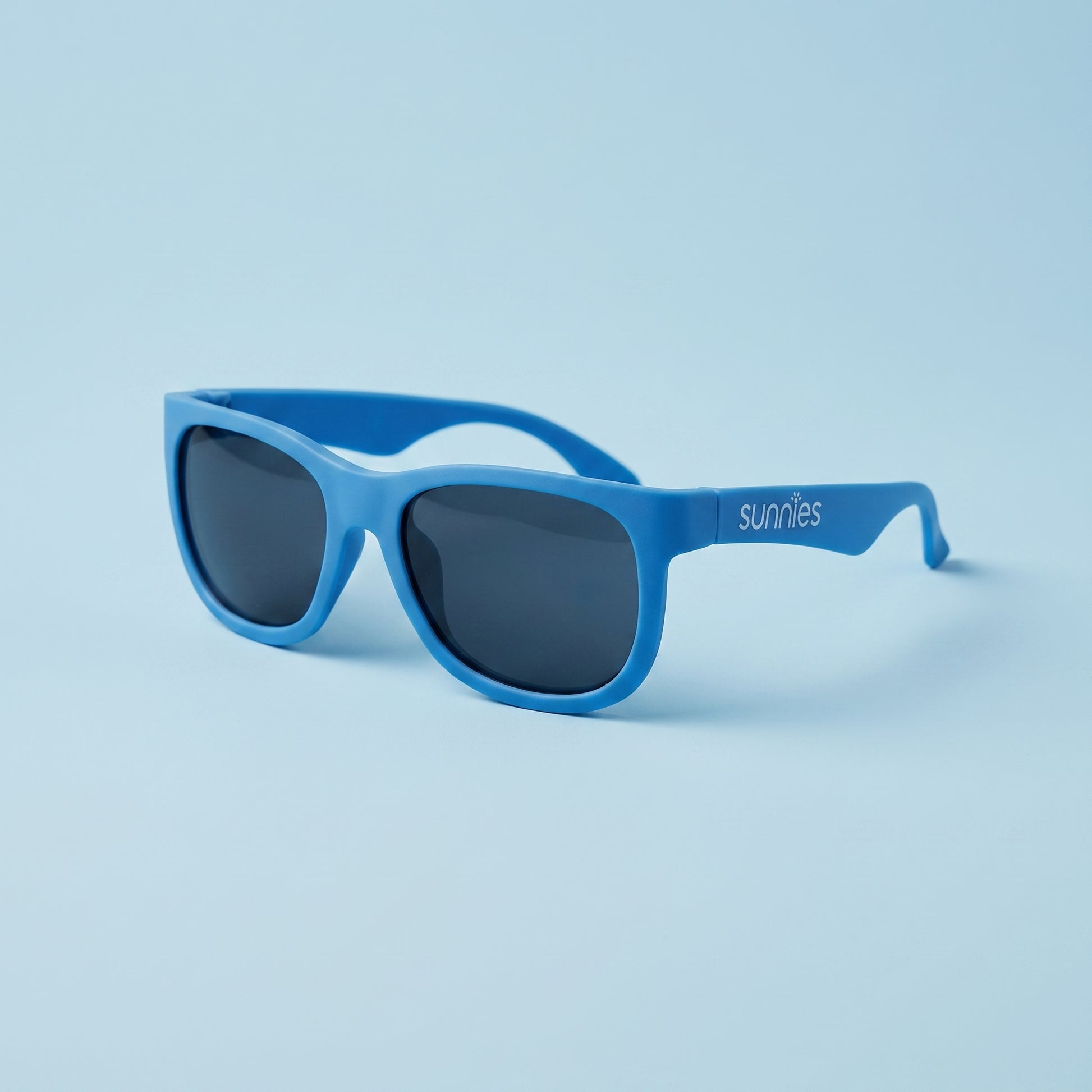hero: A NEW photorealistic photograph composed from scratch. The product, a pair of sunglasses, is centered on a seamless pastel blue surface that gently dissolves into the background. The scene is lit by broad, soft wraparound diffusion from slightly above and camera-front, complemented by gentle bounced fill that meticulously rounds the form of the product, ensuring even tonality across all surfaces. A single soft grounding contact shadow is cast directly beneath the product, anchoring it to the surface.…